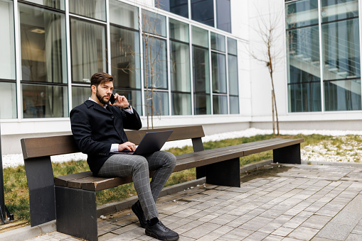 Young businessman talking on mobile phone while working on laptop outside office building
