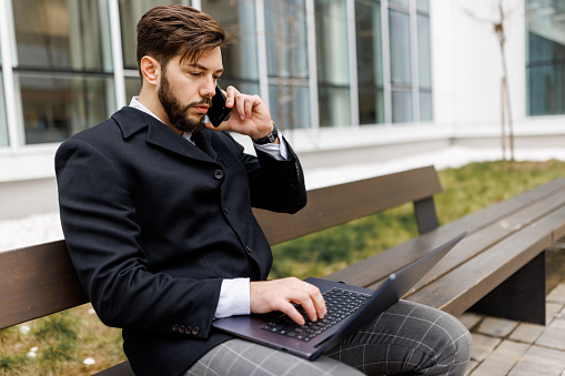 Young businessman talking on mobile phone while working on laptop outside office building