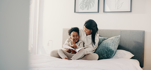 Mom and daughter laughing together as they read a story book on a bed. Young girl in elementary age is getting entertained by a fairytale she’s reading with her mother.