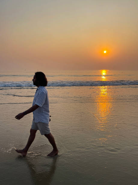 Close-up image of Indian man wearing shorts and striped short sleeved shirt holding sandals on sandy beach, vacation tourist walking barefoot at water's edge of sea at dusk, sun setting, Palolem, Goa, India, focus on foreground Stock photo showing an Indian man on holiday in Goa, South India, pictured standing on Palolem Beach, paddling in the gentle sea waves at sunset, a particularly popular winter holiday destination for both English and German tourists. palolem beach stock pictures, royalty-free photos & images