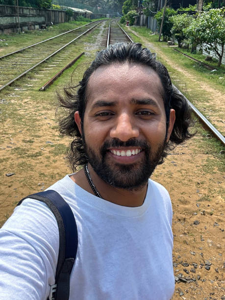 Close-up image of Indian man taking selfie with phone camera, holding mobile up and looking down at phone screen self portrait, sandy and pebbly terrain with railway train tracks background, focus on foreground stock photo