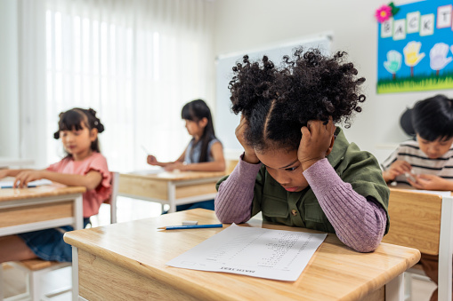 African American black girl student doing an exam at elementary school. Adorable young girl children sitting indoors on table, feeling upset and depressed while learning with teacher at kindergarten.