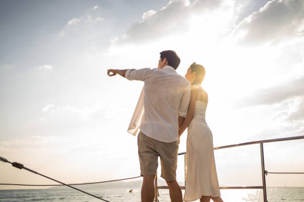 ヨット中に美しい景色を見ているアジアの若いロマンチックな夫婦。夏の日没時にカタマランボートを航行しながら、記念日の新婚旅行を祝う魅力的な男女。 - sunset yacht luxury sailboat ストックフォトと画像