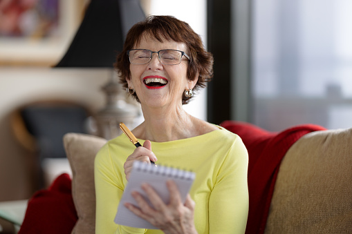 Laughing happy senior woman sitting at home with a pen and note pad.