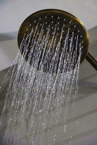 Stock photo of a round power shower in full flow, with the strong water jets being clearly visible against the bathroom tile background.  The fixed shower head cannot be adjusted, being made from brass, and appearing both stylish and expensive.