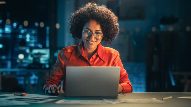 giovane donna nera che lavora al computer portatile in un ufficio creativo la sera. felice project manager multietnico che scrive e-mail, ricerca dei dettagli del piano di progetto online. - usare il laptop foto e immagini stock