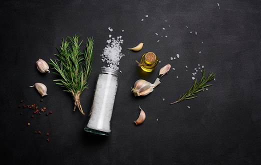 Scattered coarse white salt, peppercorns and rosemary sprigs on a black table, ingredients for cooking fish and meat
