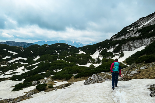 Woman with backpack crossing a snow field on a hiking trail with view on cloud covered mountain peaks of the Hochschwab Region in Upper Styria, Austria. Plateau full of snow in beautiful Alps, Europe.