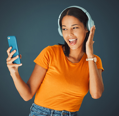 Selfie, headphones and streaming podcast or music on phone or mobile app isolated against a studio background. Fun, sound and female enjoying and listening to radio or audio smiling and happy