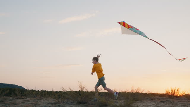 Silhouette child with kite run. School break. Lifestyle. Happy boy runs bright kite into sky, playing with wind in field sandy shore of sea an orange sunset on day lens flares in summer slow motion.