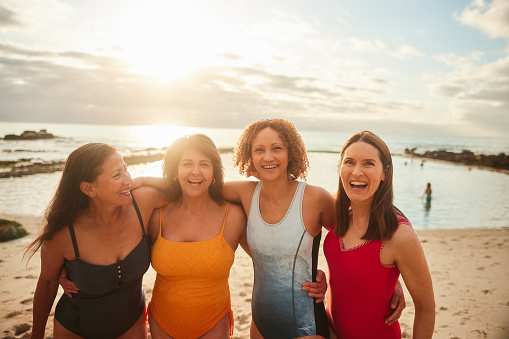 Portrait of a mature group of laughing female friends in swimwear standing arm in arm together on a sandy beach