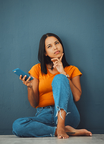 Phone, thinking and social media with a woman in studio on a blue background, sitting on the floor. Mobile, contact or idea and an attractive young female posing with her finger on her chin