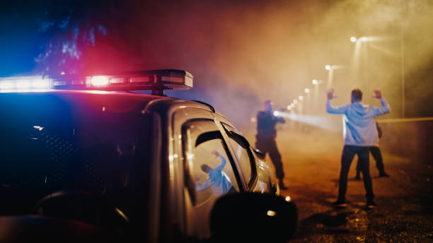 istock Two Police Officers Aim at a Fellon. Cops Using a Weapon to intimidate the Suspect. Fellon Raising Hands in Compliance. Law Enforcement Arresting a Dangerous Man. Camera Focus on Police Car Siren 1480291653