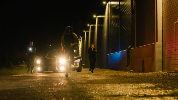 dos policías persiguiendo a un sospechoso. los policías que salen del auto comienzan a perseguir criminales a pie por las oscuras calles de la ciudad. heroicos oficiales atrapan y arrestan a traficante de drogas, asesinado - persecución fotografías e imágenes de stock