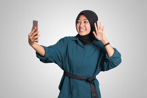 Portrait of excited Asian muslim woman with hijab holding mobile phone while in video calling with her hand gesture saying hi. Advertising concept. Isolated image on white background