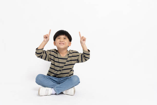 istock Happy Asian little boy pointing up with empty copy space and sitting on floor isolated on white background, Full body composition and five years old 1480292894