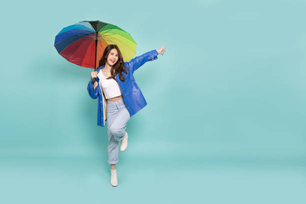 young asian woman wearing raincoat and holding rainbow umbrella isolated on green background, in the rainy season concept - rain women umbrella parasol imagens e fotografias de stock