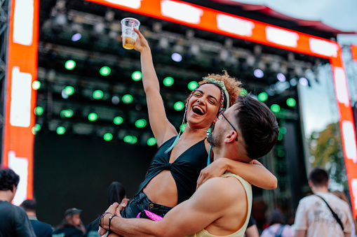 A young delighted couple is dancing at a festival. The man is holding his girlfriend up in his arms and she has a beer holding up in her hands.