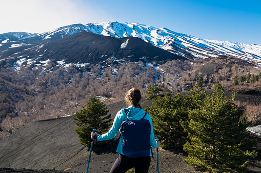 Tourist woman hiking on volcanic landscape of volcano mount Etna, in Sicily, Italy, Europe. Pine and white birch trees growing on solidified lava, ash and pumice. Slopes of crater covered with snow
