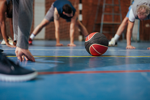 A group of people are on a basketball court stretching before playing a game with the basket ball in focus.