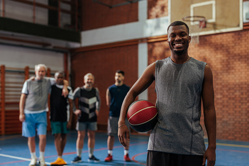 A young adult black man is standing in the gymnasium with his friends in the background after a recreational game of basketball.