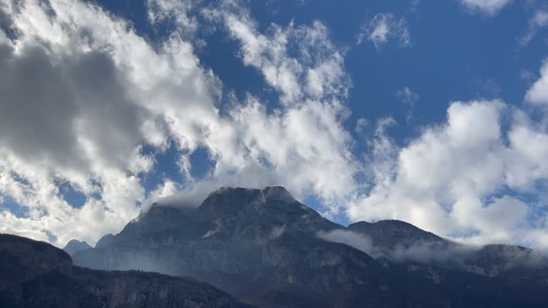 Low-hanging clouds stretch through powerful mountains against a cloudy blue sky