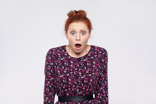 Portrait of shocked beautiful young adult redhead woman wearing dress standing, looking forward with surprised unbelievable face. Indoor studio shot isolated on gray background.