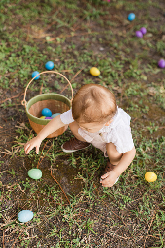 A Cute Cuban-American 15-Month-Old Baby Boy with Light Brown Hair & Brown Eyes Wearing Tan Bunny & Carrot Overalls & Brown Shoes While Holding an Easter Egg Basket, Enjoying His First Easter Egg Hunt to Celebrate the Easter Holiday in 2023. Neutral Natural Earthy Tones.
