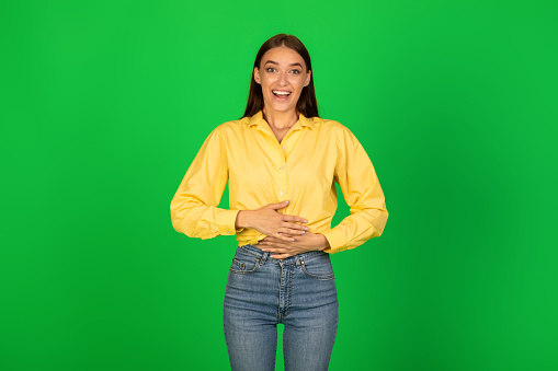 Pleasant Feeling In Stomach. Satisfied smiling young woman keeping both hands on belly, being in good mood after eating delicious meal, demonstrating that she is full, green studio background wall