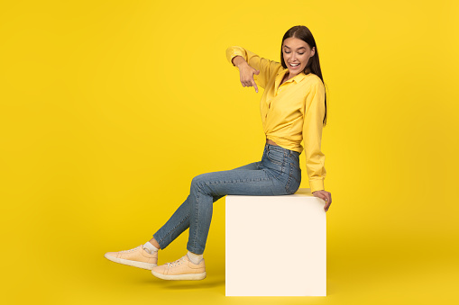 Cheerful Lady Sitting On Cube Pointing Finger Down Posing Smiling To Camera On Yellow Studio Background. Happy Female Advertising Great Offer. Full Length