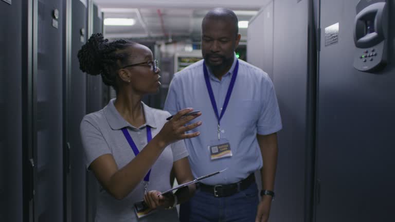 Man, woman and walking in server room, IT and maintenance of industrial computer programming. Black people, engineering and talking in data center of digital network, clipboard and cybersecurity team