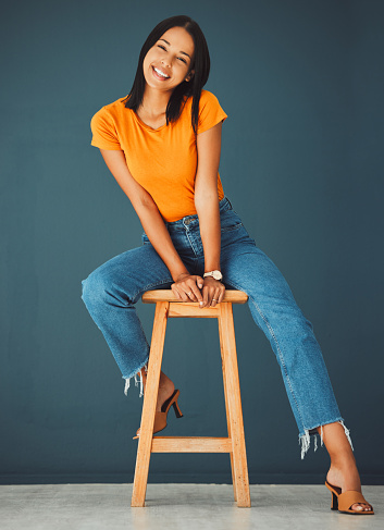 Black woman, smile and portrait of a young model sitting on a stool in a studio. Casual fashion, happiness and youth of a female feeling relax, calm and happy on a chair isolated by a blue wall