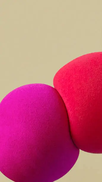 Macro shot of two spherical makeup sponges - magenta and orange, squeezing against beige background.