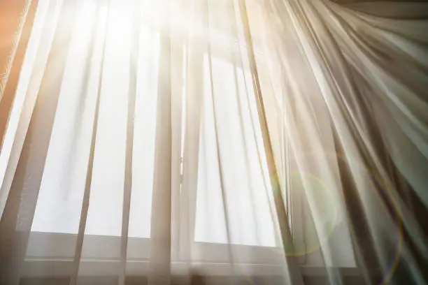 Bright sun rays lighting in house room through thin white curtain on warm summer day.