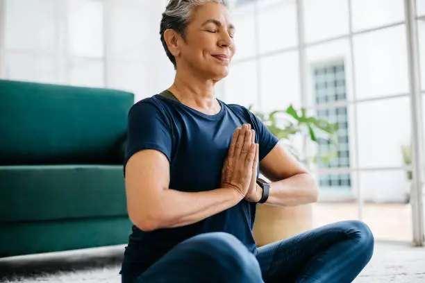 Mature woman practicing yoga meditation at home, she sits in in prayer pose with her eyes closed, breathing calmly. Senior woman doing her daily workout routine for mental and physical wellbeing.