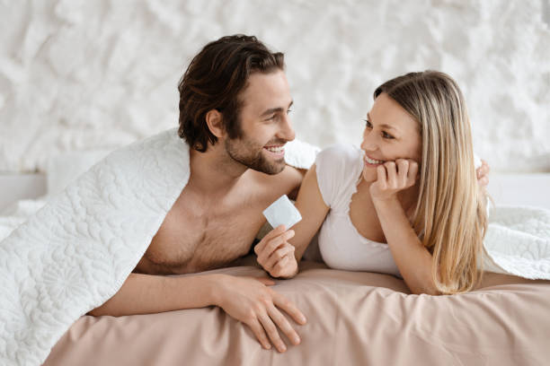 ベッドに横たわり、コンドームを持つ幸せな愛する夫婦、安全なセックスを選ぶ恋人、避妊を使う、コピー用スペース - condom contraceptive sensuality healthcare and medicine ストックフォトと画像