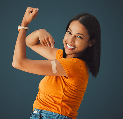 Vaccine, plaster and portrait of a woman in a studio with a strength gesture after being vaccinated. Happy, smile and proud female model with a vaccination band aid isolated by a dark blue background