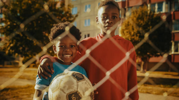 Portrait of Two Young Handsome Black Boys Embracing Each Other while Looking at Camera and Smiling. Younger Brother Holding a Soccer Ball. African Kids Standing in Urban Backyard Together.