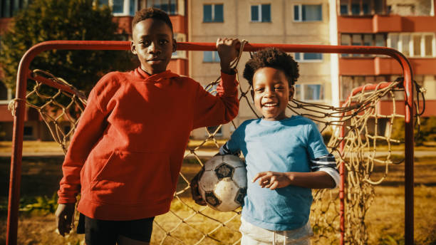 istock Portrait of Two Young Handsome Black Boys Standing Together while Looking at Camera and Smiling. Younger Brother Holding a Soccer Ball. African Kids Standing in Front of a Football Gate. 1480319635