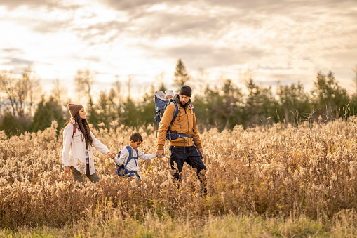 A young family of four hike their way through the tall grass on a cool fall day.  They are each dressed warmly in layers as they explore in nature together.