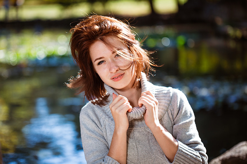 Portrait of young woman of caucasian appearance posing in sweater in an autumn park on a sunny day. A red-haired girl holds the neck of a sweater with her hands and looks at the camera