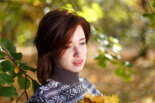 Outdoor portrait of young caucasian woman in knitted sweater posing against nature background. A beautiful woman with a pensive expression is resting in the park. Against the background of trees