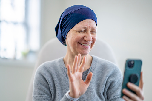 A middle aged woman who is battling cancer, sits in the comfort of her own home as she video chats with family on her cell phone.  She is dressed comfortably and has a headscarf on to keep her warm as she smiles and chats with family.