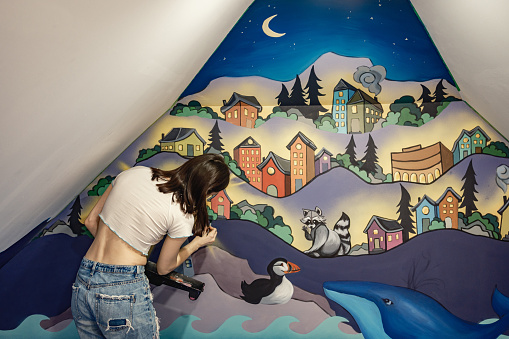 Young female artist painting mural in the attic. She is dressed in casual work clothes with ripped jeans. Interior of private home in North America.