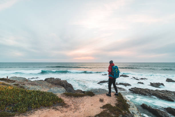 Fearless man with a backpack watching the sunset over the Atlantic Ocean in Porto Covo, Portugal. A hiker stands on the edge of a cliff looking into his future. Self-awareness stock photo