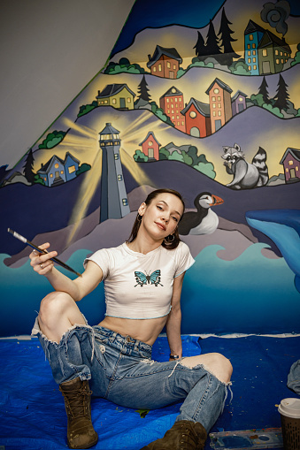 Location Portrait of Young female artist painting mural in the attic. She is dressed in casual work clothes with ripped jeans. Interior of private home in North America.