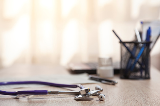Close-up of a stethoscope over a wooden table where are, out of focus, a pens pot, paper, medicines and a laptop. There are a window on background.