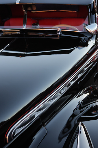 Vintage classic car in shiny black finish with red pinstripe and red interior - in a tightly cropped close-up vertical composition that captures portions of the hood, front quarter panel and fender, windshield and interior from an angle - camera positioned above the front driver's side headlight looking downward toward the hood.  The primary image color theme is black, with a hint of red in a pinstripe along the front quarter panel and in the interior.  Chrome accents also appear at several points.  No people appear in the photo.