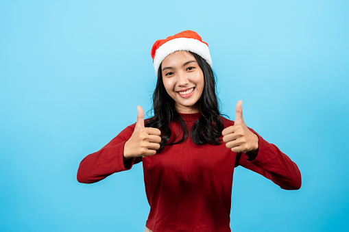Portrait. Girl with long hair in a red tights wearing a Christmas hat. Raise one hand with a trembling thumb and extend it forward. It's a great gesture. Indoor studio isolated on blue background