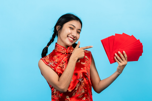 portrait, girl wearing cheongsam on chinese new year, Count red envelopes in your hand one by one, and raises envelope show camera, smiling, Isolated indoor studio on blue background